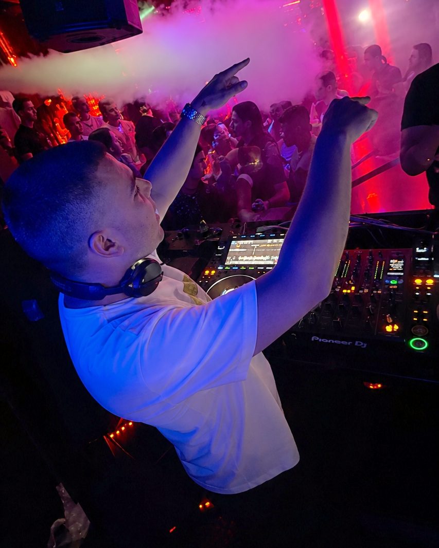 DJ Orestis with both arms raised behind the decks, crowd and laser lighting behind him