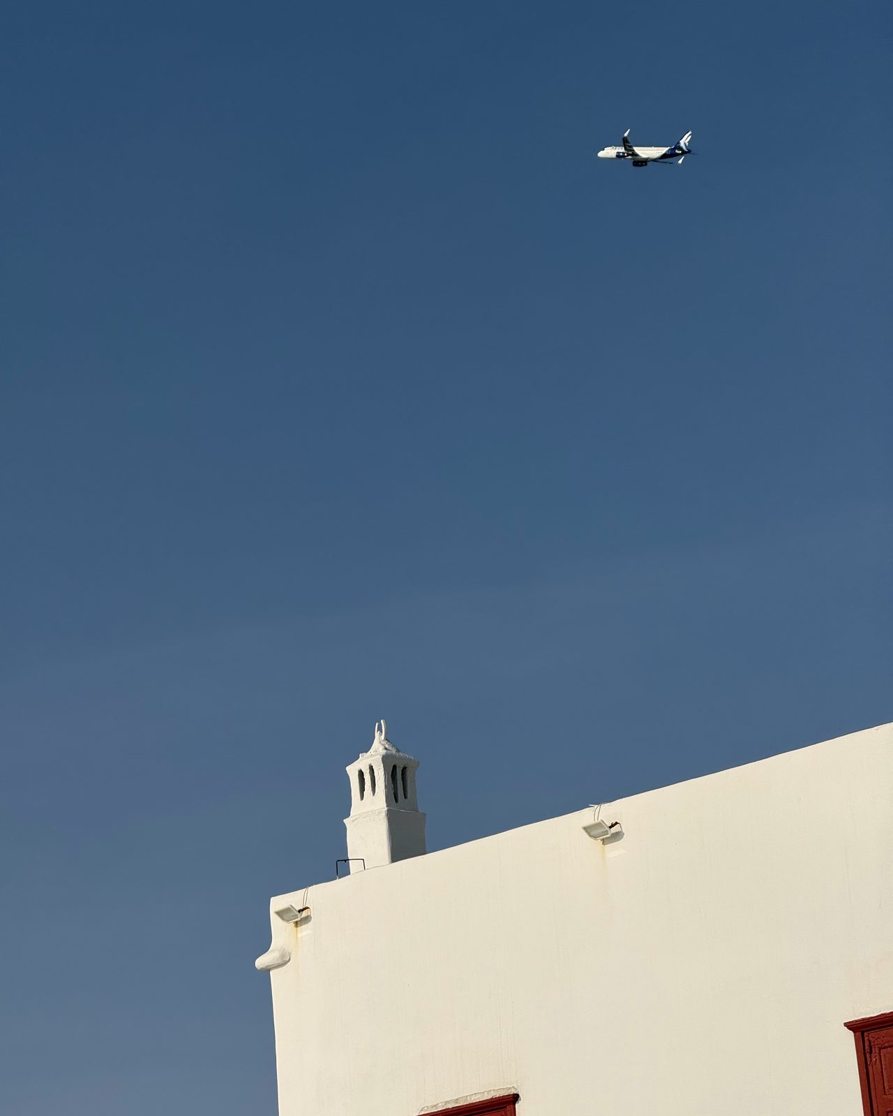 A whitewashed Mykonos building with a red door and a plane crossing the blue sky above